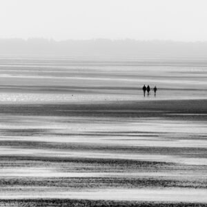 Das Wandbild Ruhig bei Ebbe zeigt eine weite Küstenlandschaft in Schwarz-Weiß, in der drei Personen in der Ferne über den feuchten Meeresboden spazieren.