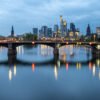 Das Wandbild Skyline Frankfurt zeigt die beleuchtete Bankenmetropole mit der Ignatz-Bubis-Brücke und Spiegelungen im Main zur blauen Stunde.