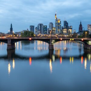 Das Wandbild Skyline Frankfurt zeigt die beleuchtete Bankenmetropole mit der Ignatz-Bubis-Brücke und Spiegelungen im Main zur blauen Stunde.