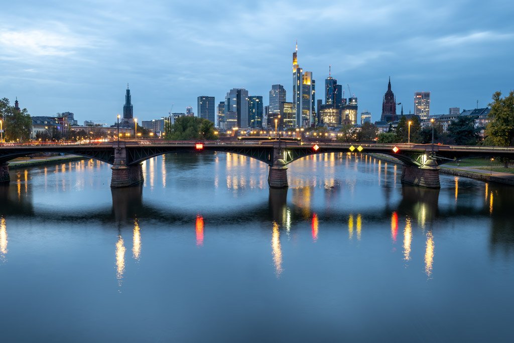 Das Wandbild Skyline Frankfurt zeigt die beleuchtete Bankenmetropole mit der Ignatz-Bubis-Brücke und Spiegelungen im Main zur blauen Stunde.