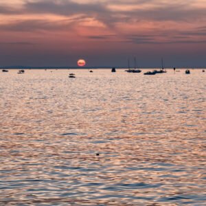 Das Wandbild Sonnenuntergang am See zeigt eine weite Wasserfläche mit vielen Segelbooten und Motorbooten unter einem rötlichen Abendhimmel, an dem die Sonne tief am Horizont steht.