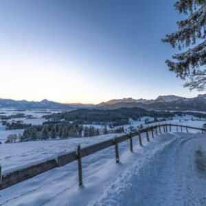 Das Wandbild Winterlicher Allgäumorgen zeigt eine weite, tief verschneite Allgäuer Landschaft bei Sonnenaufgang mit einem hölzernen Zaun im Vordergrund und den markanten Alpengipfeln am Horizont.