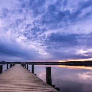 Das Wandbild Wörthsee zeigt einen langen Holzsteg, der mittig in den spiegelglatten See führt, unter einem dramatischen Wolkenhimmel in Violett- und Goldtönen während der Dämmerung.