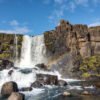 Das Wandbild Öxarárfoss zeigt den isländischen Wasserfall mit einem leuchtenden Regenbogen vor den dunklen Basaltfelsen unter blauem Himmel