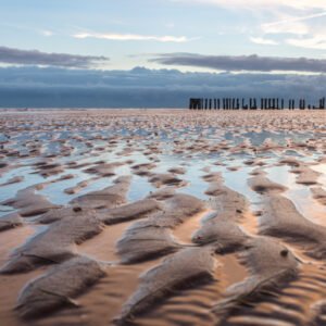 Das Wandbild Watt voraus zeigt die charakteristischen Wellenstrukturen im Sand bei Ebbe unter einem weiten Himmel mit einer Holzpfahl-Reihe am Horizont