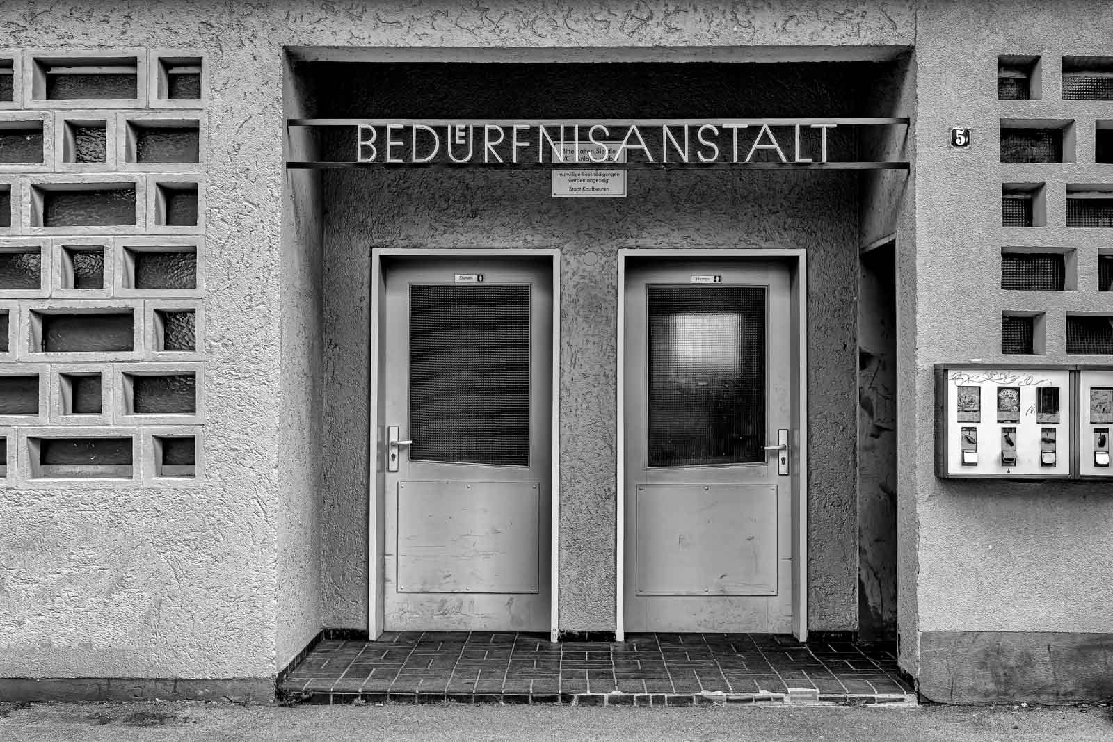 Symmetrische Frontalaufnahme einer historischen Bedürfnisanstalt mit zwei Türen in Schwarz-Weiß.