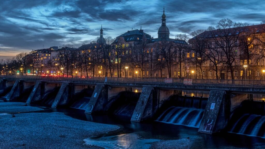 Das Isarwehr in München während der blauen Stunde mit beleuchteten historischen Gebäuden im Hintergrund.