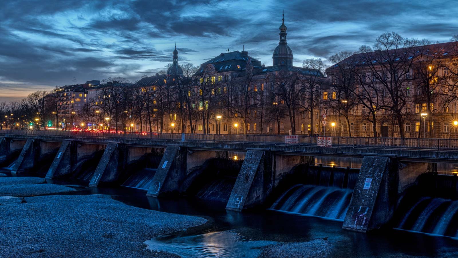 Das Isarwehr in München während der blauen Stunde mit beleuchteten historischen Gebäuden im Hintergrund.