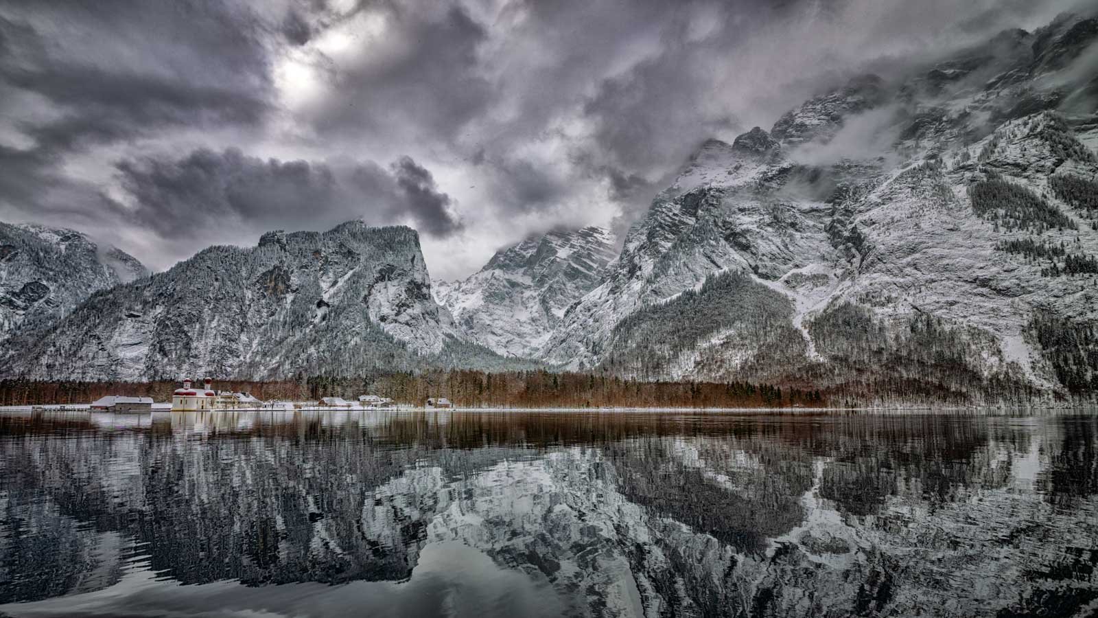 Dramatische Winterlandschaft am Königssee mit St. Bartholomä vor verschneiten Bergmassiven und bewölktem Himmel.