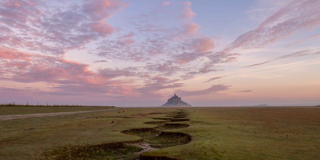 Le Mont Saint Michel am Horizont bei Sonnenaufgang mit rosa Wolken – Fotografie Roland Seichter.