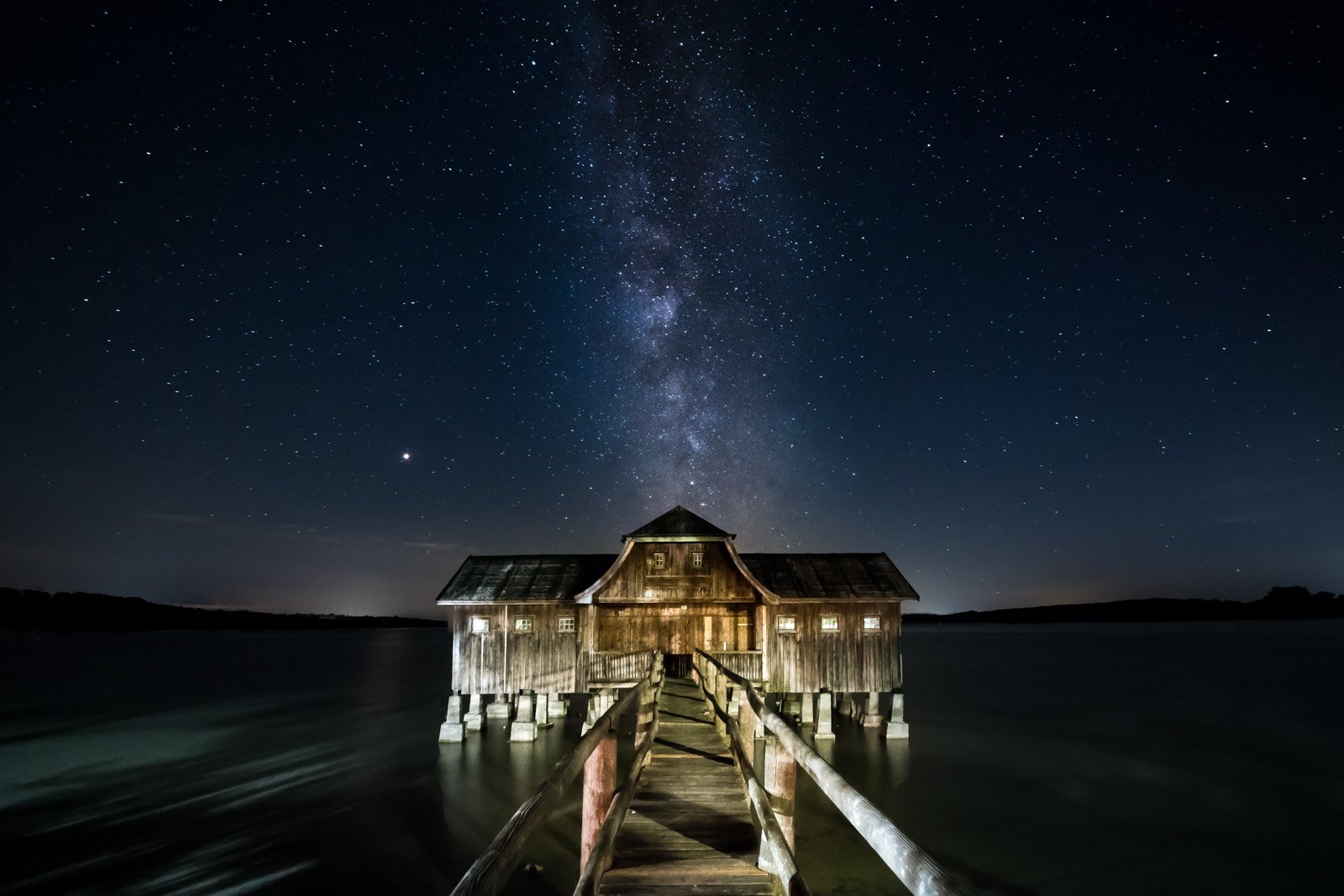 Die Milchstraße über einem beleuchteten Bootshaus am See bei Nacht.