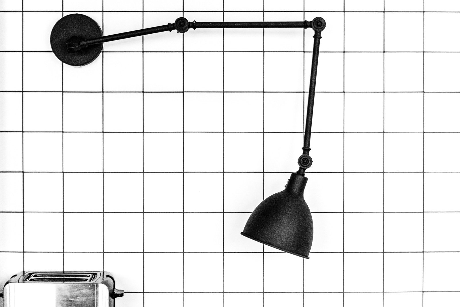 Minimalistische Schwarz-Weiß-Aufnahme einer Gelenklampe vor einer weißen Kachelwand mit Toaster.