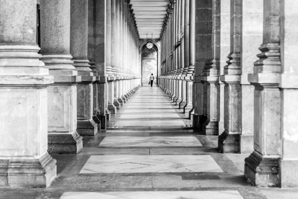 Schwarz-weiß Fotografie eines langen, symmetrischen Säulengangs mit einer Person in der Ferne und einer großen Uhr.