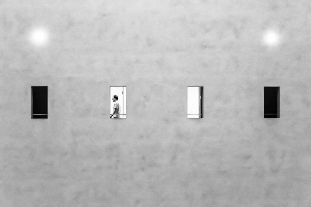 Minimalistische graue Fassade mit vier kleinen Fenstern, in einem ist die Silhouette eines Mannes sichtbar.