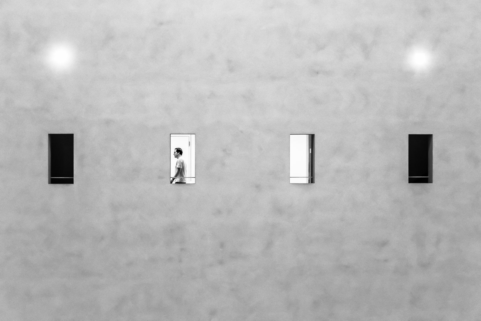 Minimalistische graue Fassade mit vier kleinen Fenstern, in einem ist die Silhouette eines Mannes sichtbar.