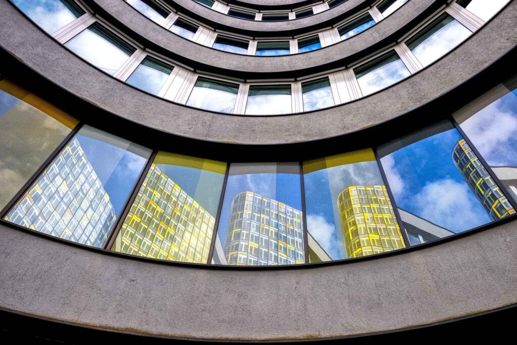 Moderne Architekturfassade mit geschwungenen Fensterfronten, in denen sich gelbe Gebäude und der blaue Himmel spiegeln.