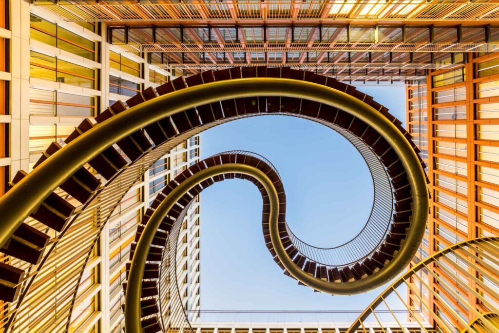 Blick von unten auf eine geschwungene, endlose Stahltreppe vor einem modernen Bürogebäude und blauem Himmel.