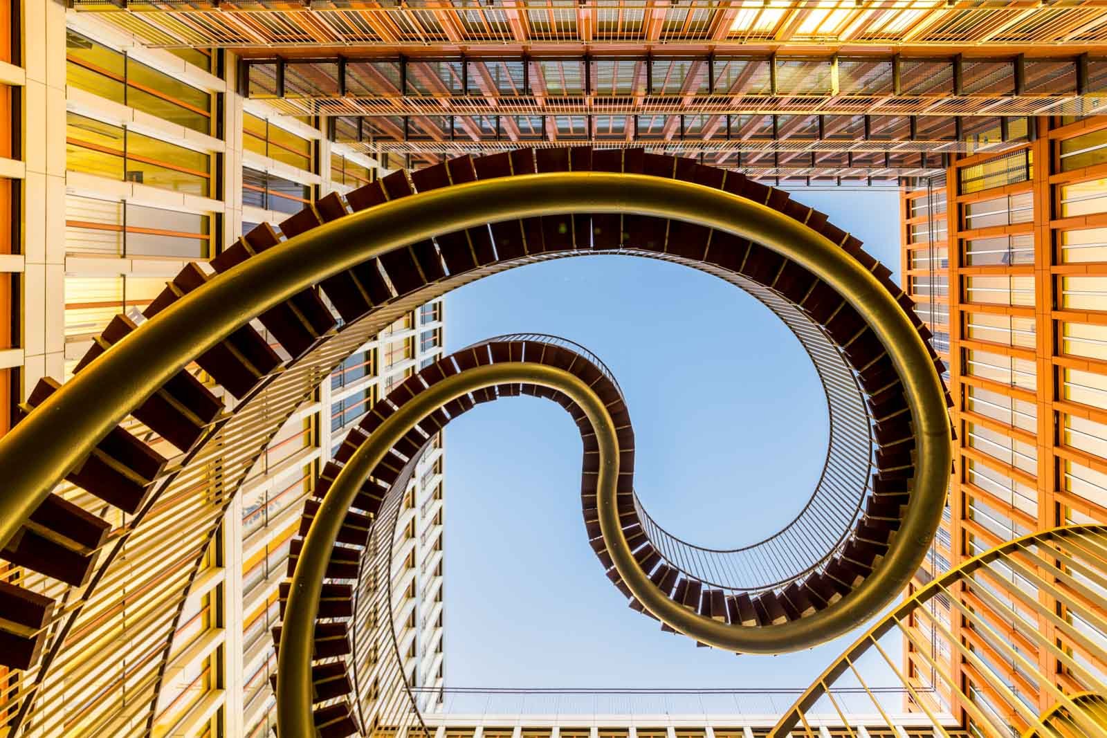 Blick von unten auf eine geschwungene, endlose Stahltreppe vor einem modernen Bürogebäude und blauem Himmel.