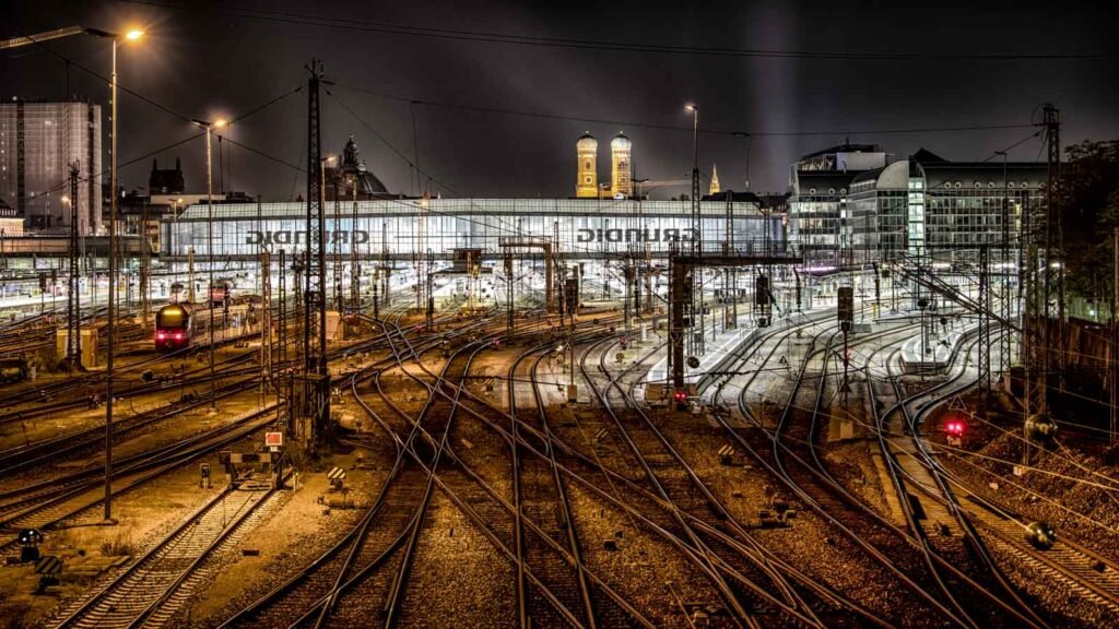 Nachtaufnahme der Gleisanlagen am Münchener Hauptbahnhof mit der Frauenkirche im Hintergrund.
