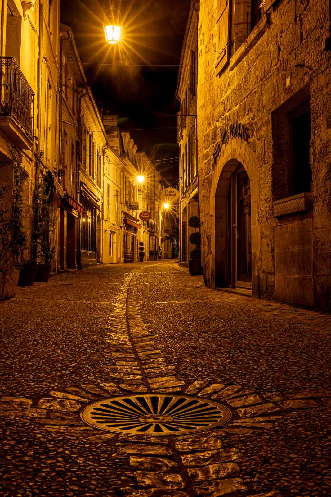 Eine stimmungsvoll beleuchtete Altstadtgasse bei Nacht mit Pflastersteinen und einer markanten Kanalabdeckung.