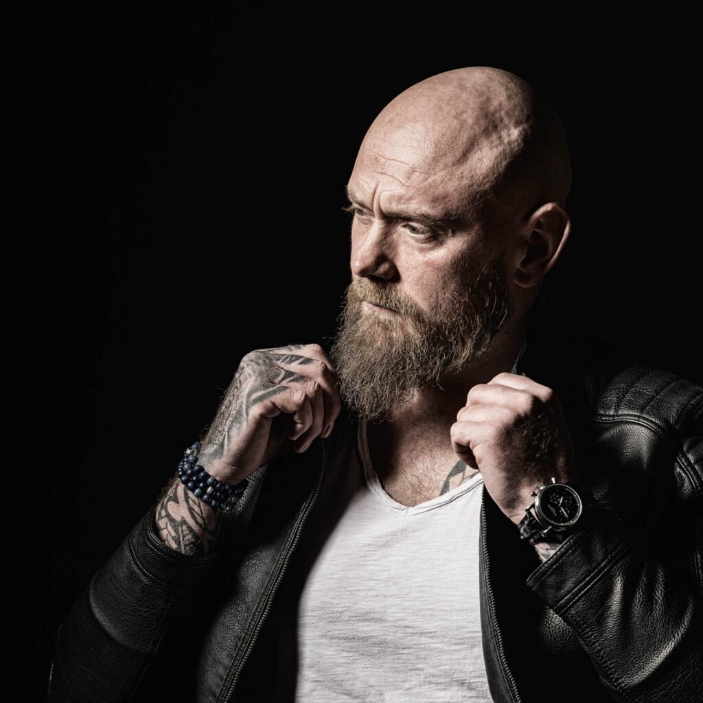 Porträt eines bärtigen Mannes mit Glatze in Lederjacke, der nach links blickt und seine Jackenkragen hält.