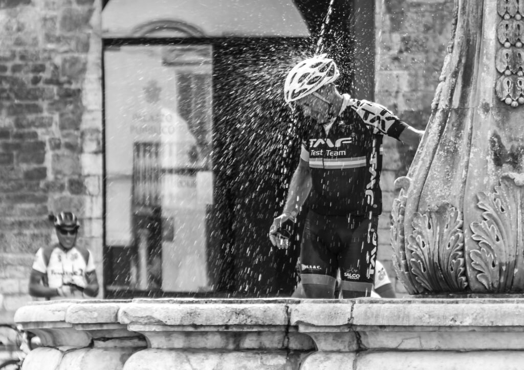 Ein Radrennfahrer kühlt sich unter dem Wasserstrahl eines historischen Brunnens ab.