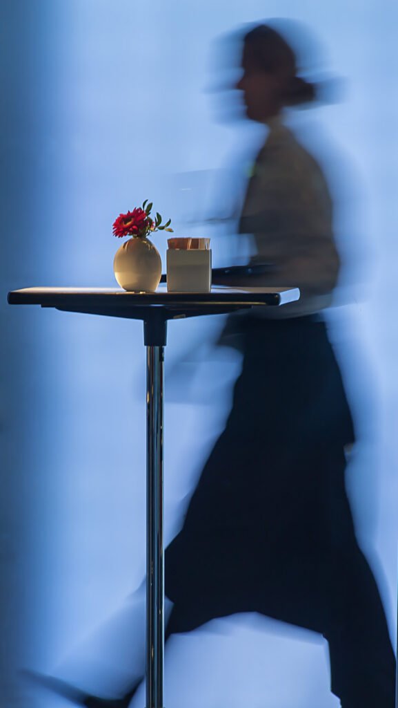 Eine scharf fokussierte rote Blume in einer Vase auf einem Stehtisch, im Hintergrund eine Person in dynamischer Bewegungsunschärfe.