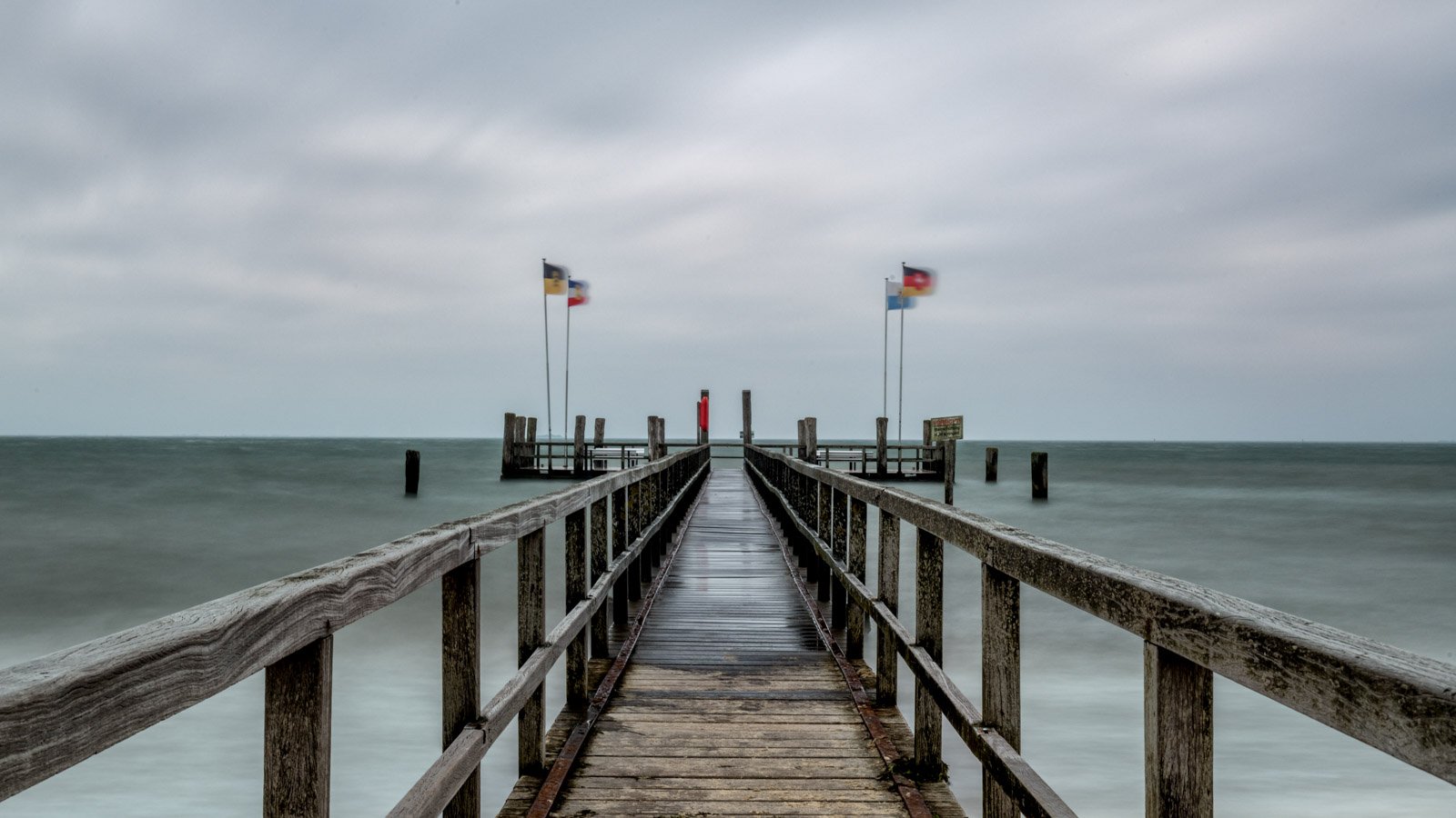 Symmetrische Aufnahme eines Holzstegs an der Nordsee unter bewölktem Himmel.
