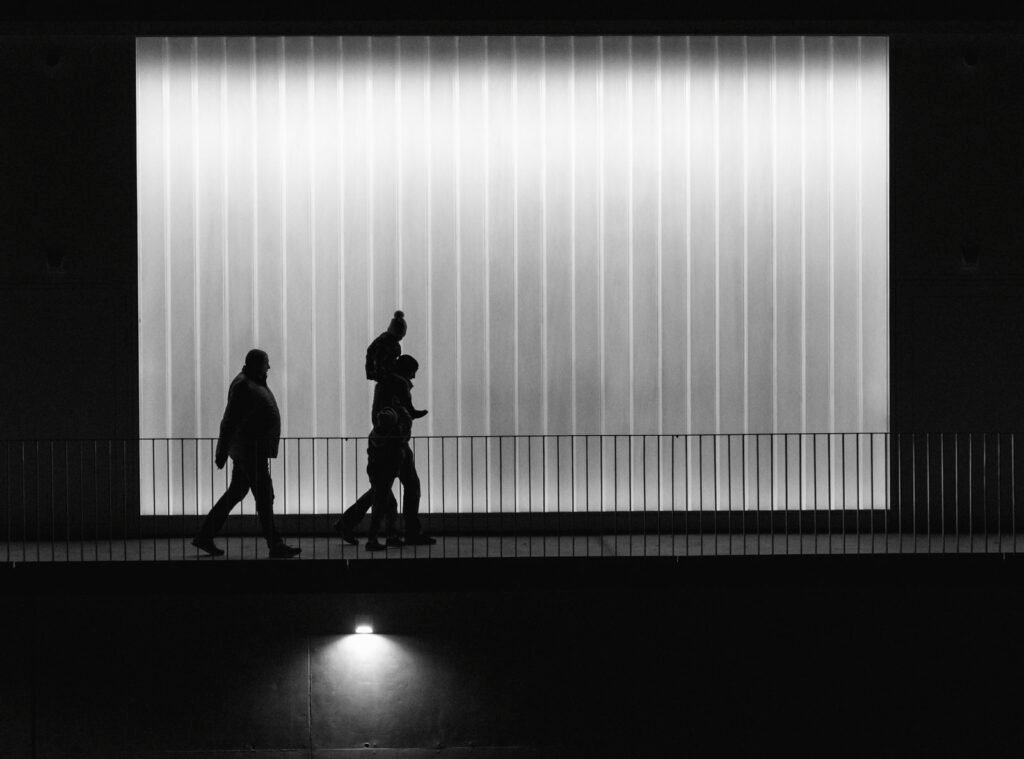 Silhouetten von vorbeigehenden Menschen vor einer hell leuchtenden Glaswand in moderner Architektur.