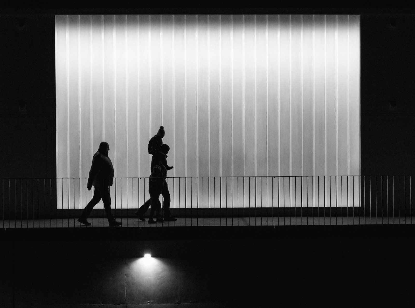 Silhouetten von vorbeigehenden Menschen vor einer hell leuchtenden Glaswand in moderner Architektur.