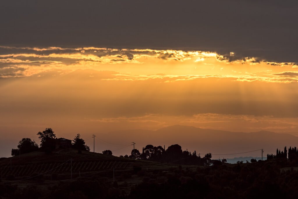 Silhouette eines toskanischen Gehöfts vor einem dramatischen, gold-orange leuchtenden Sonnenuntergang.
