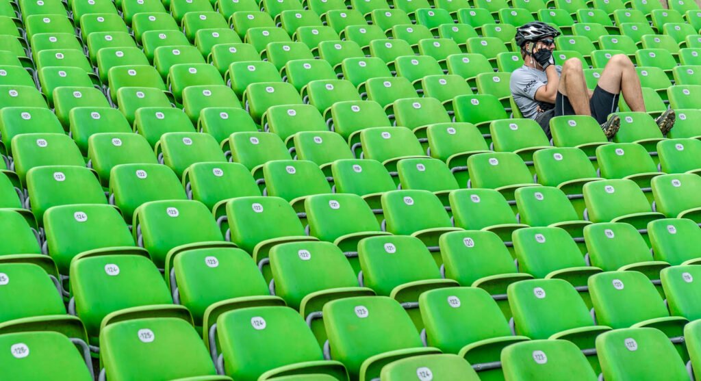 Ein Radfahrer mit Helm und Handy am Ohr rastet in den leeren grünen Sitzreihen eines Stadions.