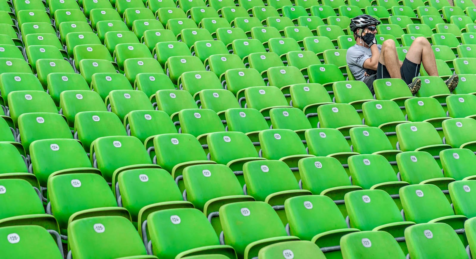 Ein Radfahrer mit Helm und Handy am Ohr rastet in den leeren grünen Sitzreihen eines Stadions.