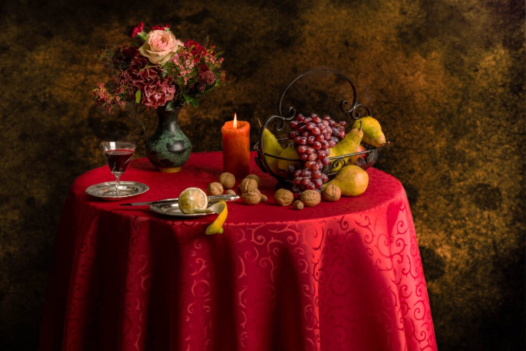 Klassisches Stillleben mit Obstschale, Blumenstrauß und Wein auf einer roten Tischdecke.