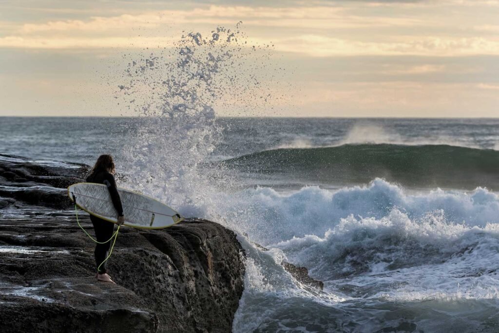 Ein Surfer steht mit seinem Brett an einer felsigen Küste und beobachtet die aufpeitschende Gischt der Wellen.