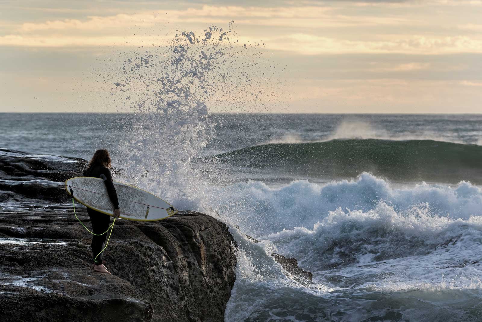 Ein Surfer steht mit seinem Brett an einer felsigen Küste und beobachtet die aufpeitschende Gischt der Wellen.