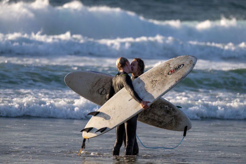 Ein Surfer-Paar küsst sich im flachen Wasser am Strand vor brandenden Wellen.