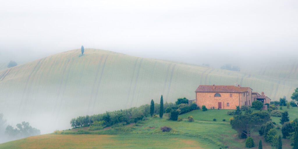 Ein steinernes Landhaus in der Toskana vor einem nebelverhangenen Hügel mit Zypressen.