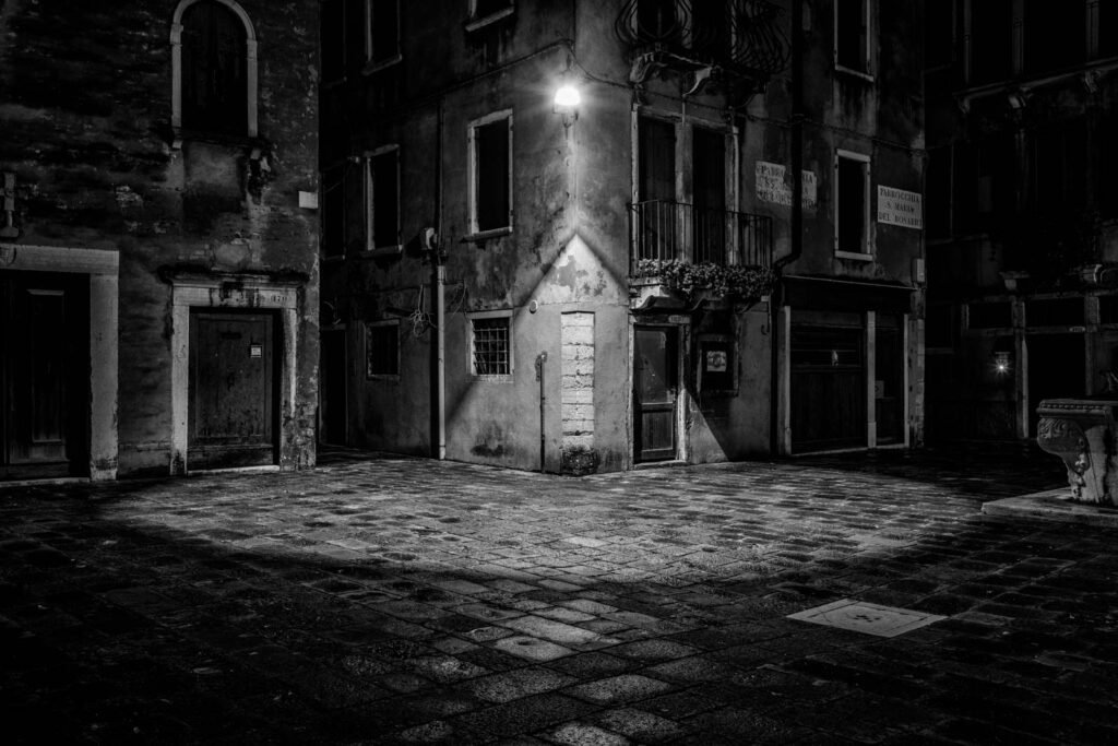 Nächtliche Szene in einer Gasse in Venedig mit einem markanten Lichtkegel einer Straßenlaterne in Schwarz-Weiß.