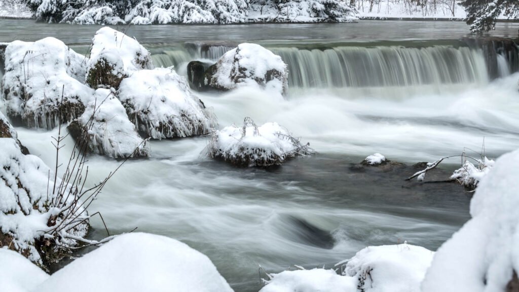 Langzeitbelichtung eines Wasserfalls im Winter mit verschneiten Steinen und fließendem Wasser.