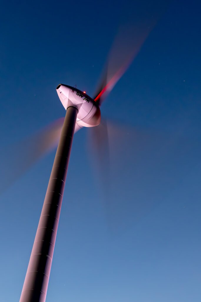 Eine moderne Windkraftanlage bei Nacht mit durch Langzeitbelichtung verschwommenen Rotorblättern vor einem dunkelblauen Sternenhimmel.