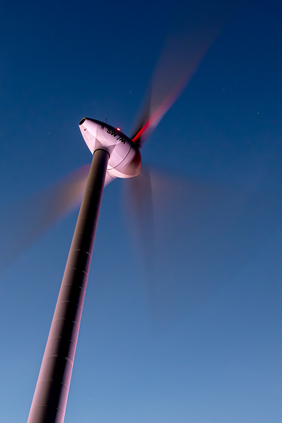 Eine moderne Windkraftanlage bei Nacht mit durch Langzeitbelichtung verschwommenen Rotorblättern vor einem dunkelblauen Sternenhimmel.