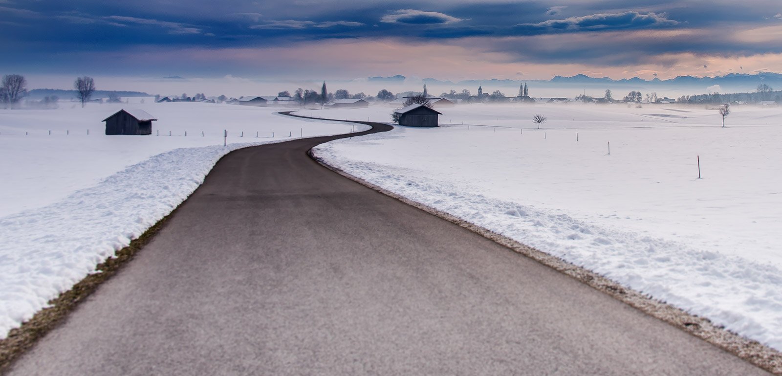 Eine einsame, geschwungene Straße führt durch eine verschneite Landschaft mit Alpenpanorama.
