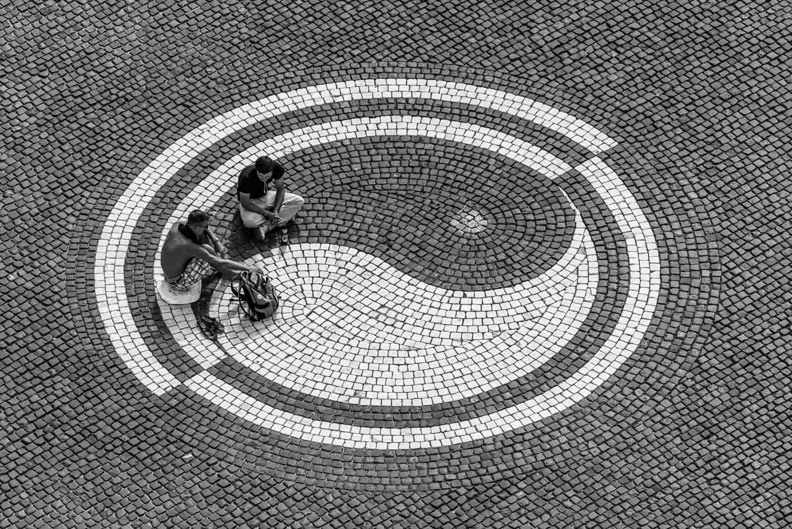 Draufsicht auf zwei Personen, die in einem in das Kopfsteinpflaster eingelassenen Yin-Yang-Symbol sitzen.